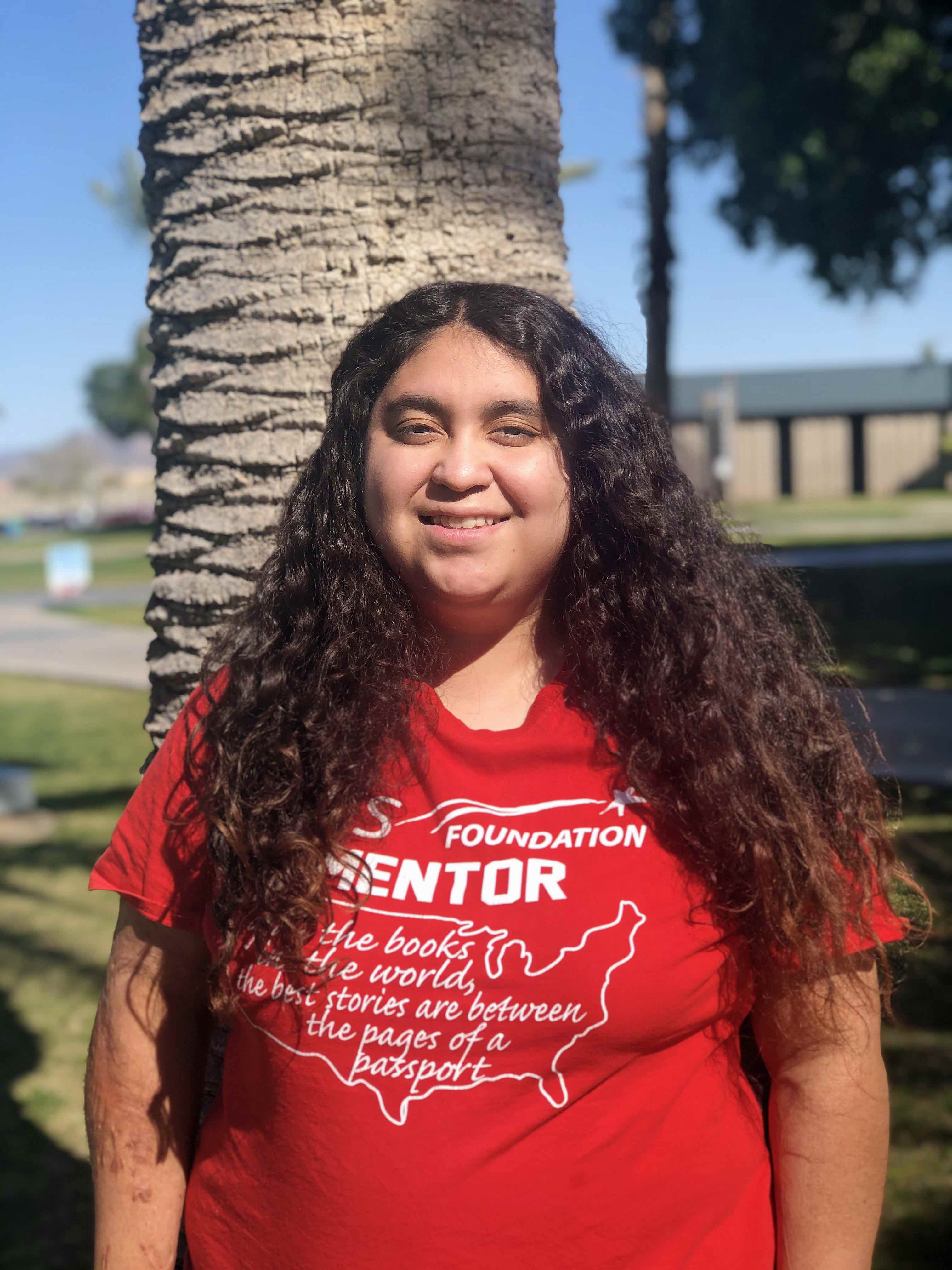 A girl with long black curly hair is wearing a red shirt that says "Foundation Mentor" and is smiling in front of a palm tree.