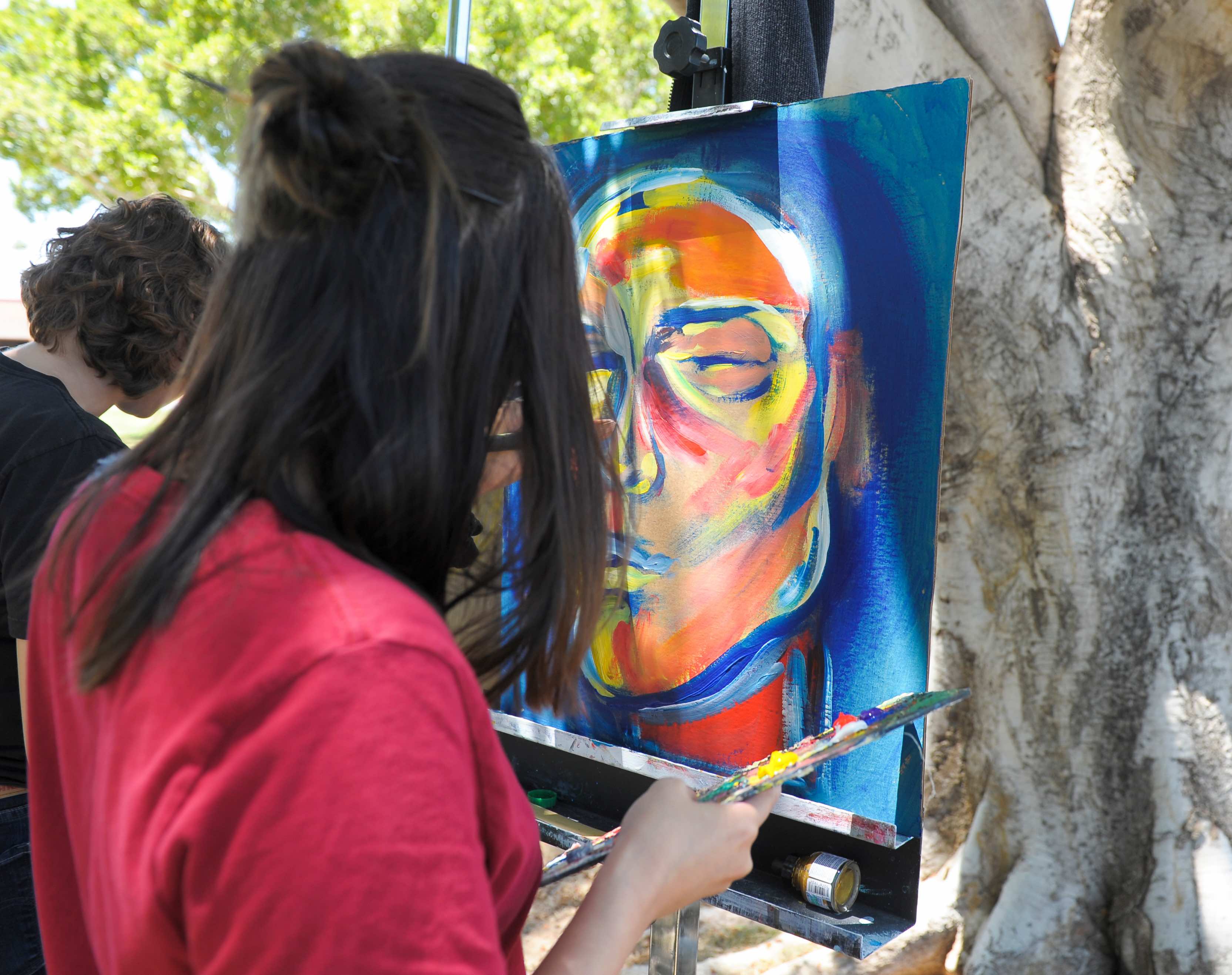 A person in red is standing outside painting on a canvas which is already blue with an orange face 