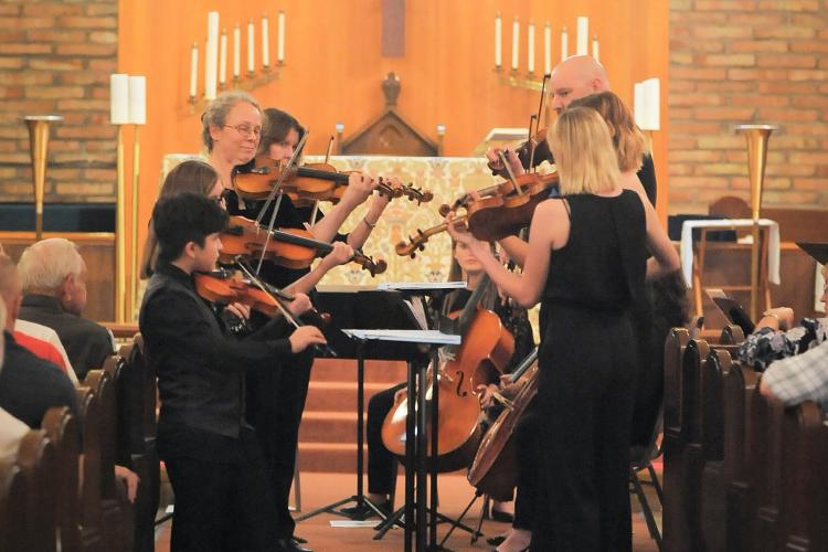String Ensemble director Margaret Fenske (3rd from left, facing camera) performs during a Nov. 2017 concert. Photo by Daniel Puentes, AWC Photo Services. Performers stand playing string instruments in a church